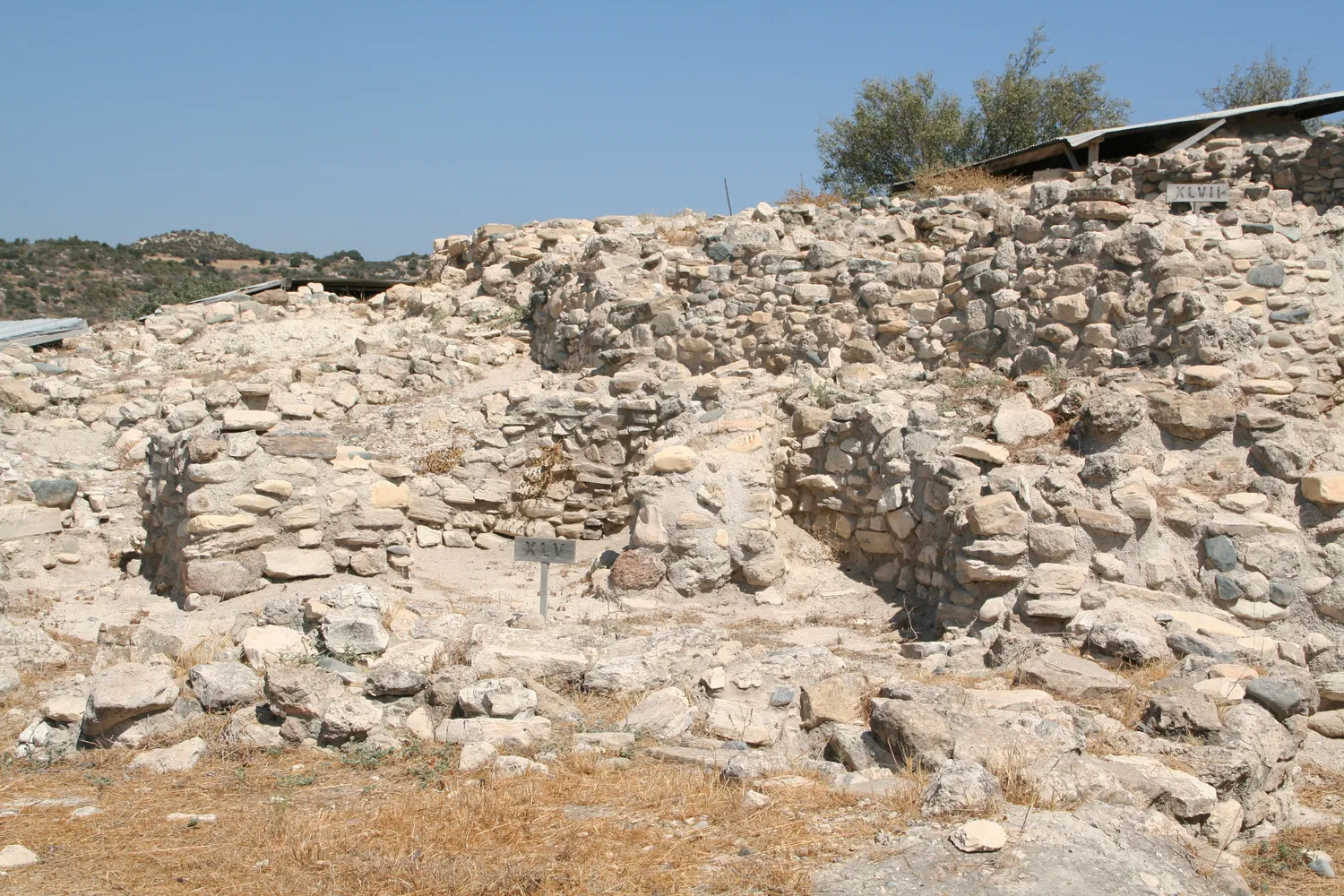 The stone walls of Tholos XLV (small sign at centre) and, behind and above it, Tholos XLVII (sign at top right), the unit whose inward-curving wall Dikaios read as the springing of a dome. Photo: Ophelia2, CC BY-SA 3.0.