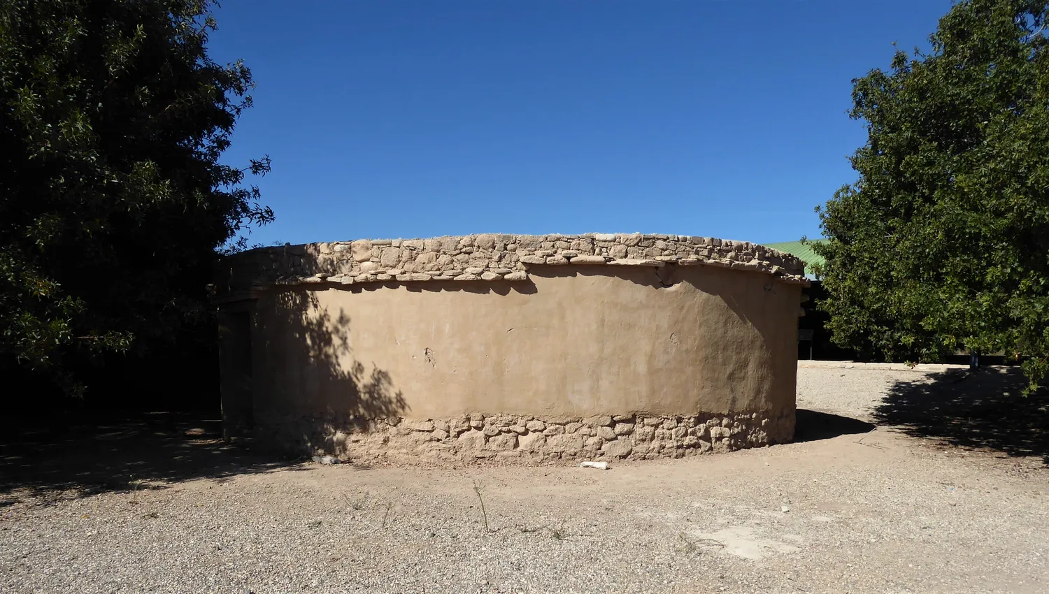 The round house rebuilt in 2010 at Kissonerga-Mosphilia on the Late Chalcolithic pattern: a smooth mud-rendered wall on a low stone footing, with a flat roof edged in stones. A modern reconstruction, not an ancient building. Photo: Chris06, CC BY-SA 4.0.