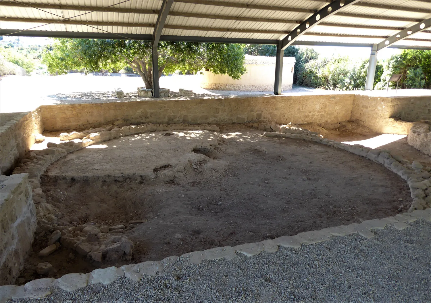 Stone footings of Chalcolithic round houses at Kissonerga-Mosphilia, displayed under a modern shelter: a ring of footing stones around a dished earth floor, with another curved wall behind. The straight walls and the roof are modern. Photo: Chris06, CC BY-SA 4.0.
