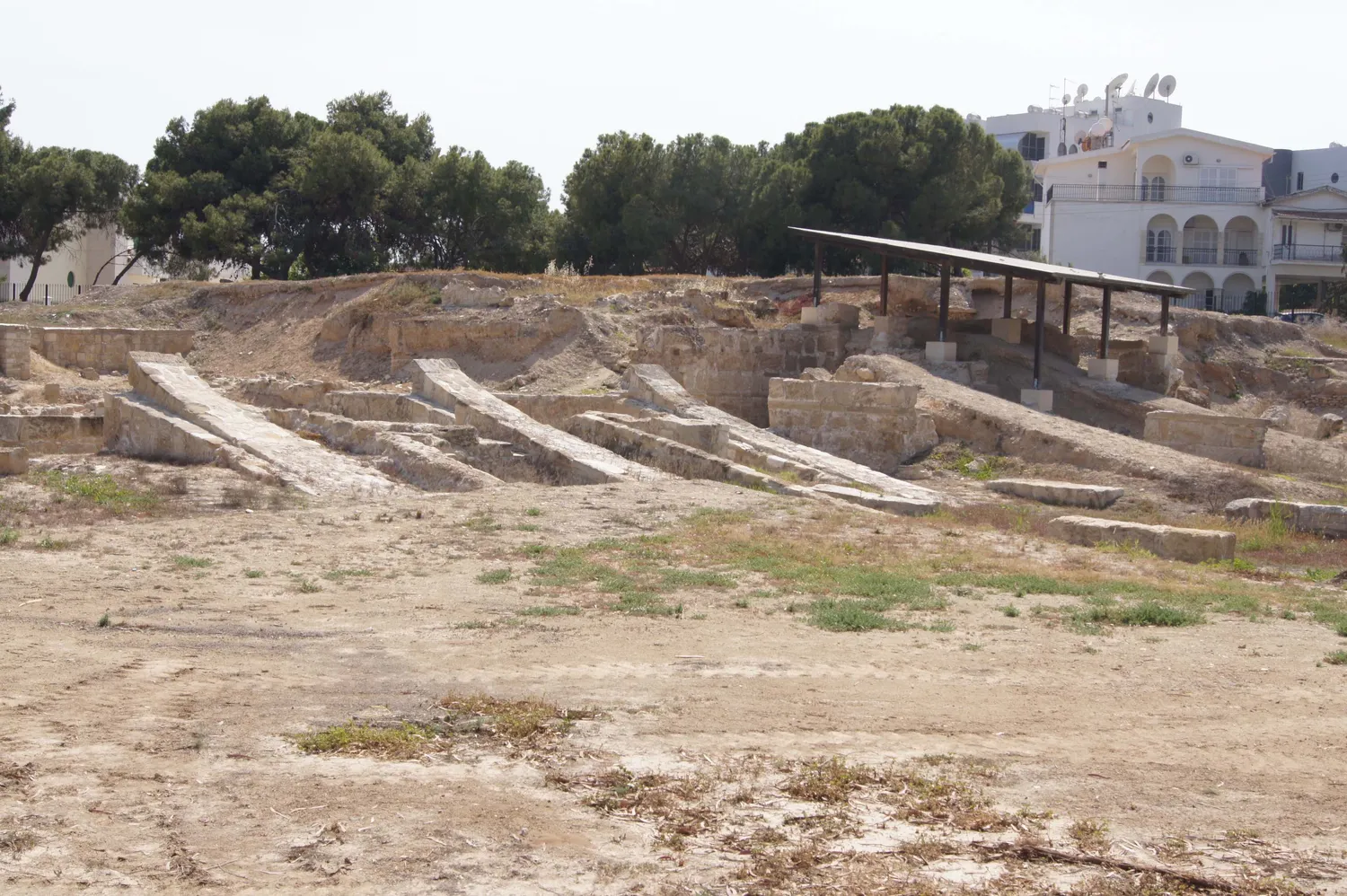 The excavated shipsheds at Kition-Bamboula in Larnaca in 2017: long stone slipways sloping down side by side towards the filled-in harbour basin, with a modern shelter roof over part of the site and town buildings behind. Photo: Rjdeadly, CC BY-SA 4.0.