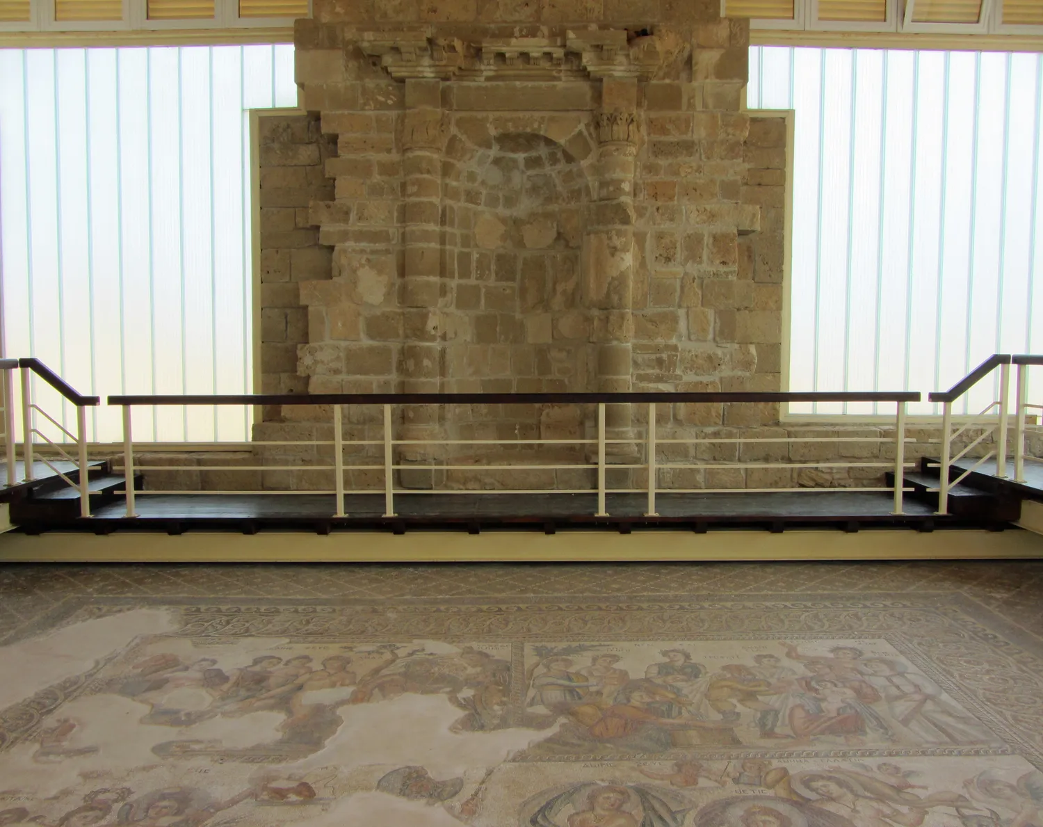 The wall of the House of Aion at Nea Paphos, found fallen in the street in 1983 and re-erected: a semicircular niche between two half-columns under a carved cornice. It stands today inside the modern shelter, behind the mosaic floor of the main hall. Photo: Stefan Bellini, CC0.