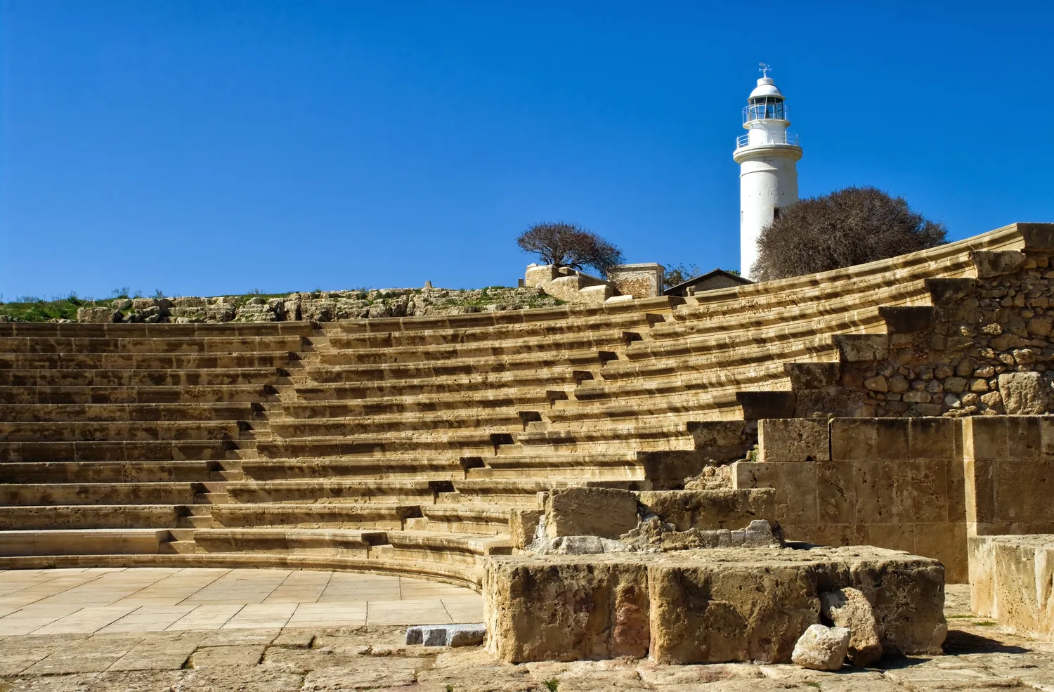 The Odeion at Nea Paphos, with the Paphos lighthouse behind it. The neat, sharp-edged rows of seats are a restoration of the early 1970s; the rougher walling at the right edge is ancient. Photo: Nikodem Nijaki, CC BY-SA 3.0.
