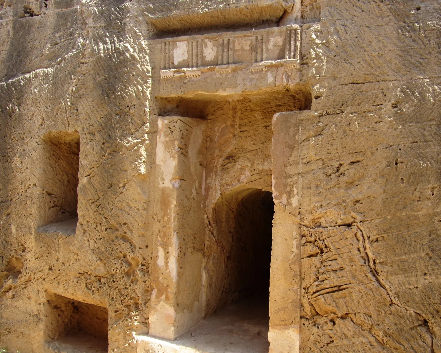 A doorway in Tomb 8 at the Tombs of the Kings, Nea Paphos, framed by two flat piers and a lintel carved with a triglyph frieze, all cut from the living rock; to the left, smaller rock-cut niches. Photo: Stefan Bellini, CC0.