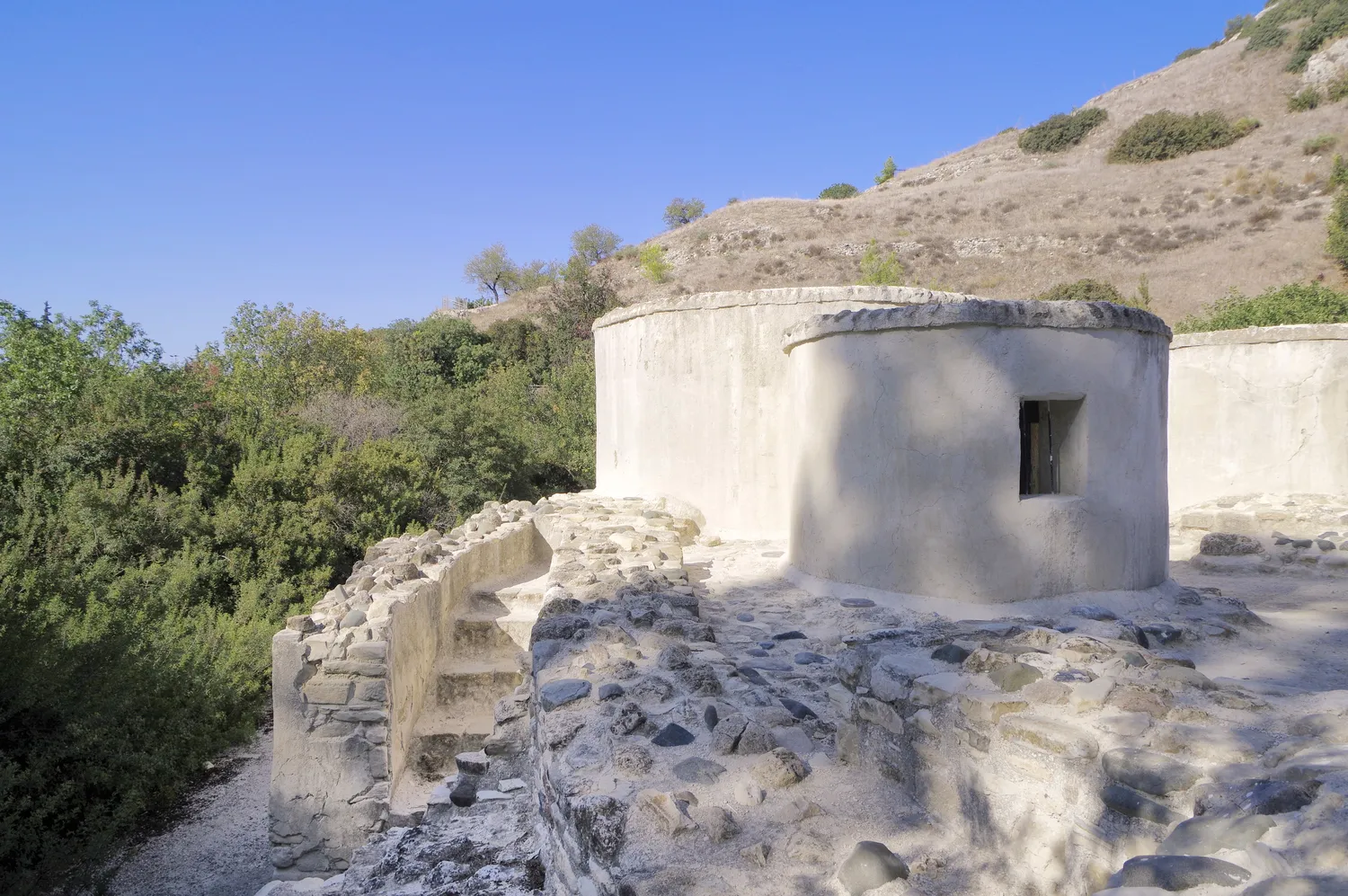 The full-size modern replica of Khirokitia's access 2 at the foot of the site: a narrow stair climbs between stone walls inside a masonry block to the top of the village wall, with replica round houses behind. Everything in the picture is modern. Photo: Berthold Werner, CC BY-SA 4.0.
