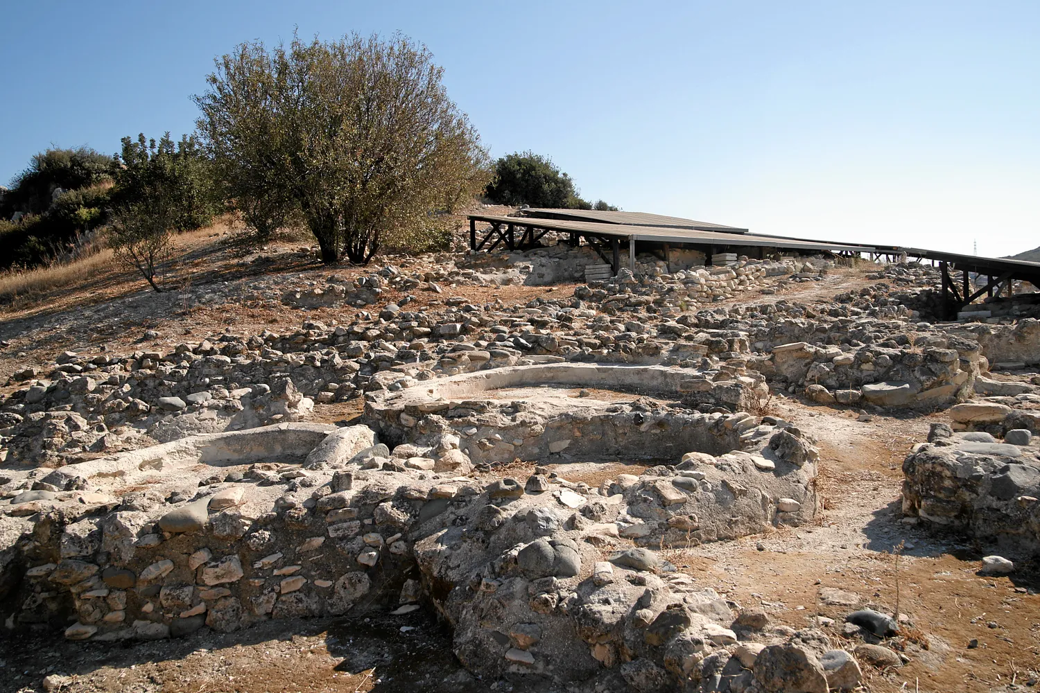 Excavated round houses at Khirokitia: low curving walls of river cobbles and limestone, one ring built against and over another, with modern shelters over the excavation behind; the smooth rims on some wall tops are modern capping. Photo: Mboesch, CC BY-SA 4.0.