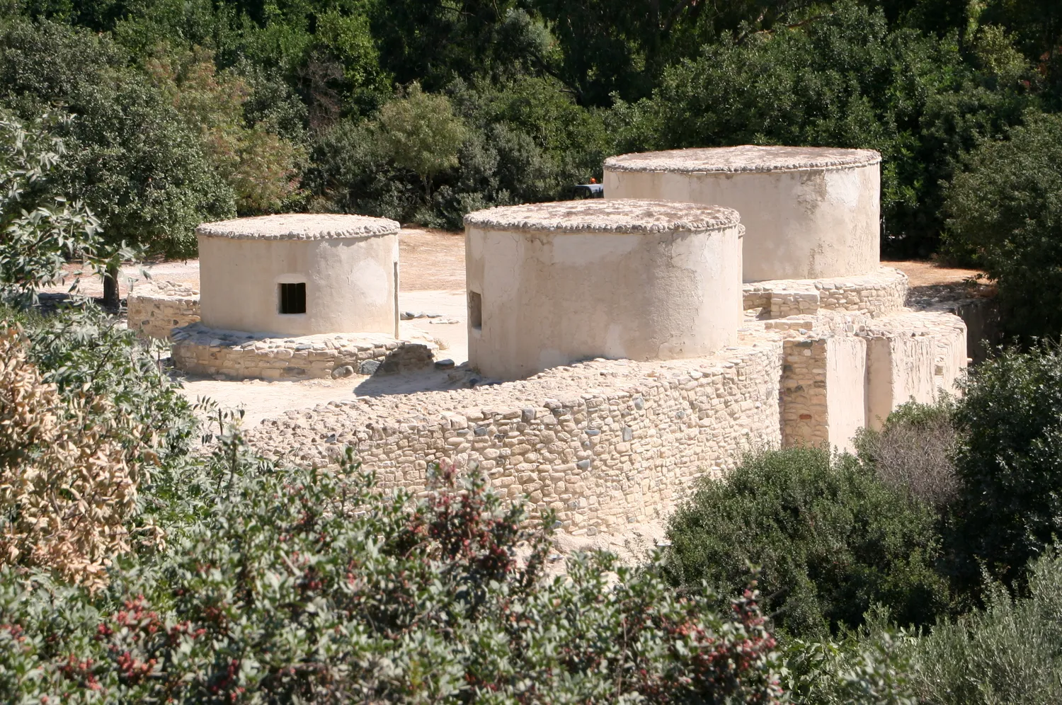 The replica houses at Khirokitia: flat-roofed cylinders of pale earth plaster standing on a stone platform among shrubs. They were built in the 1990s from the excavators' data and are modern buildings. Photo: Ophelia2, CC BY-SA 3.0.