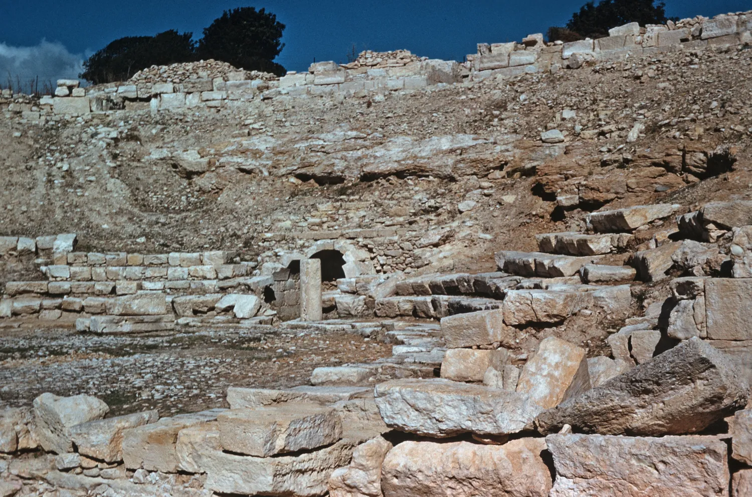The theatre at Kourion in 1958, before the seating was rebuilt: the bare, rock-cut bed of the bowl rises behind a few rows of worn seats, a small vaulted opening and a spill of tumbled blocks. Photo: Don Christie, CC BY-SA 4.0.