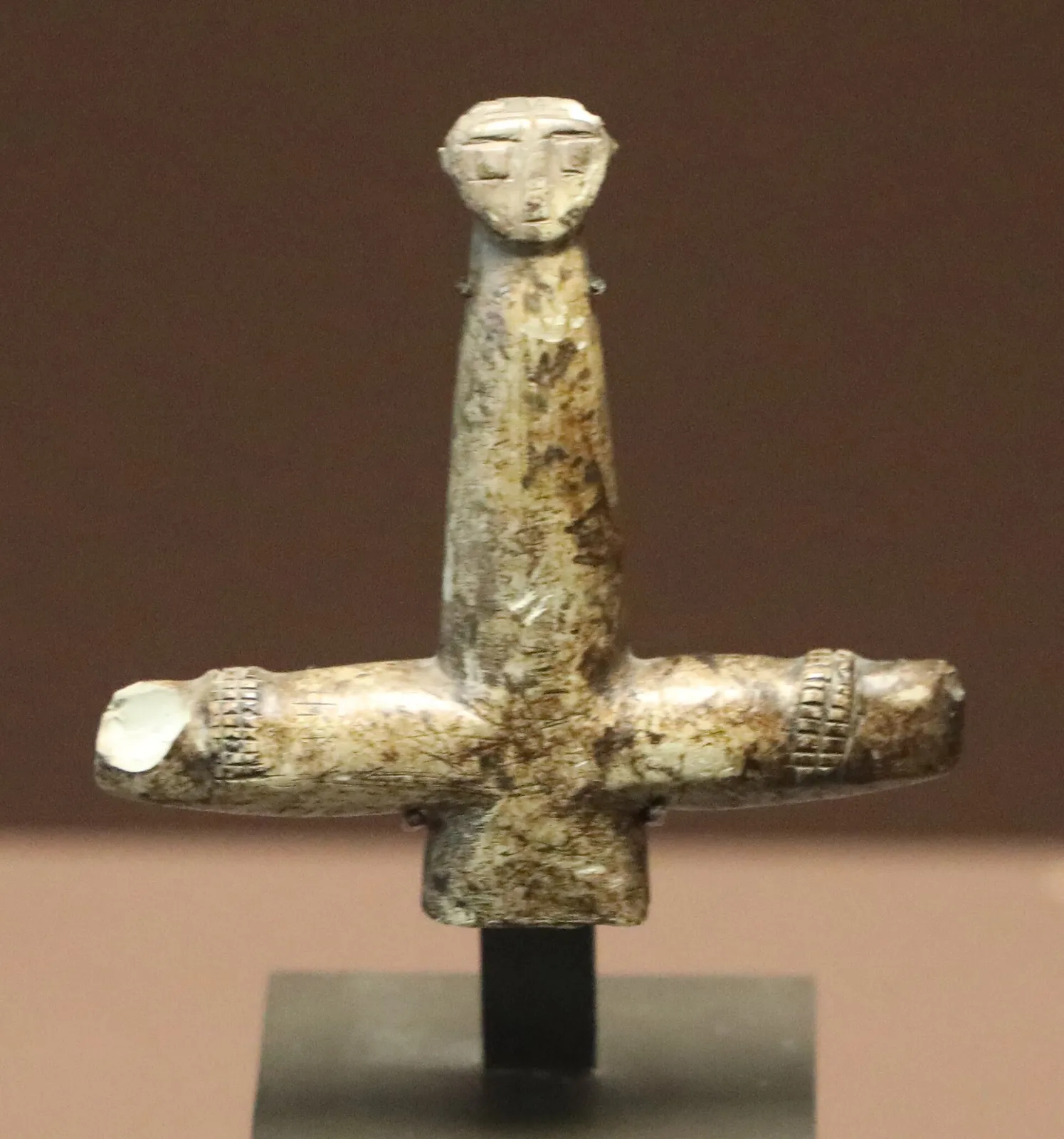 A cruciform picrolite figure with a long neck and carved face, its arms ending in notched bands, on a dark stand