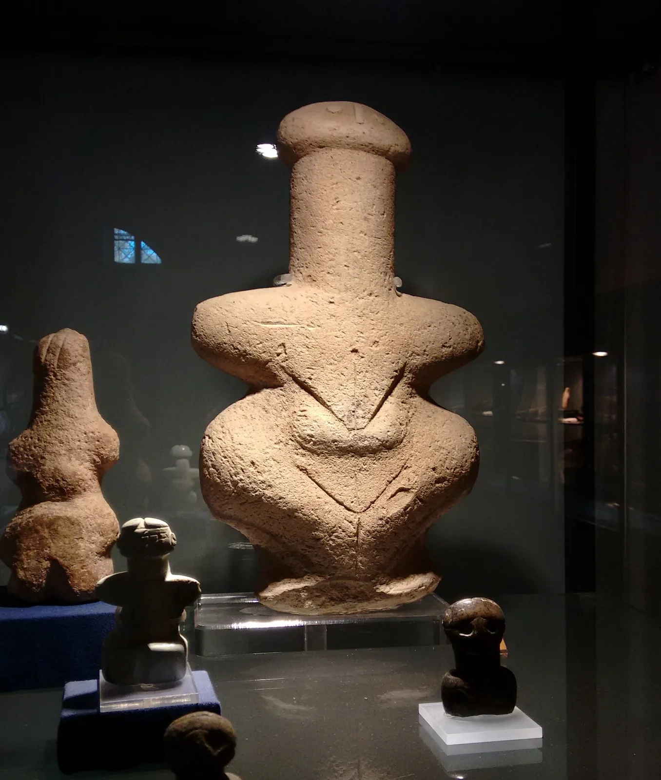 A large flat-bodied stone figure with outstretched arms, a tall head and a V-shaped incised line on the chest, in a lit museum case with smaller figures beside it