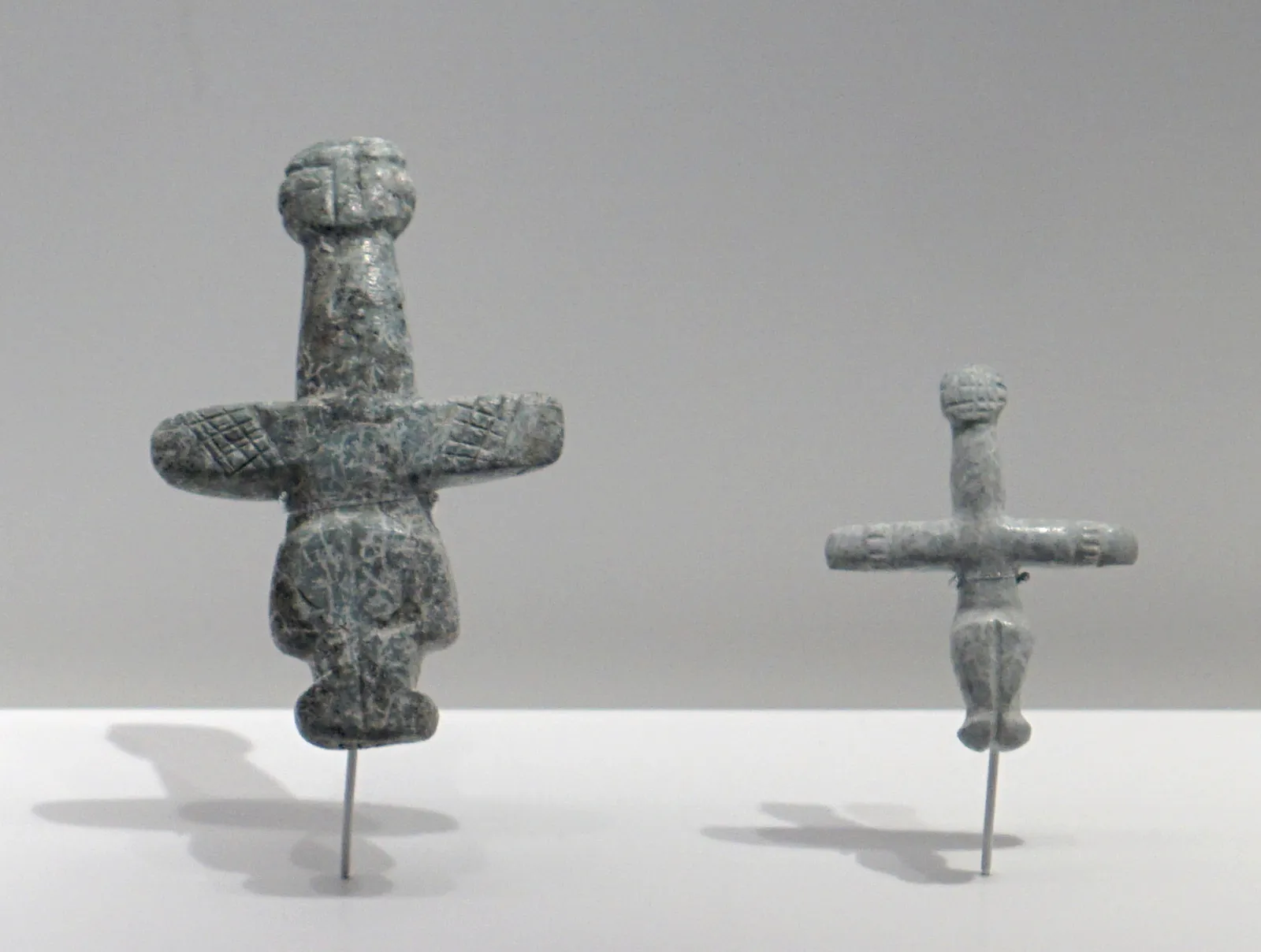 Two picrolite cruciform figures of different sizes on stands, the larger with incised lines across the body