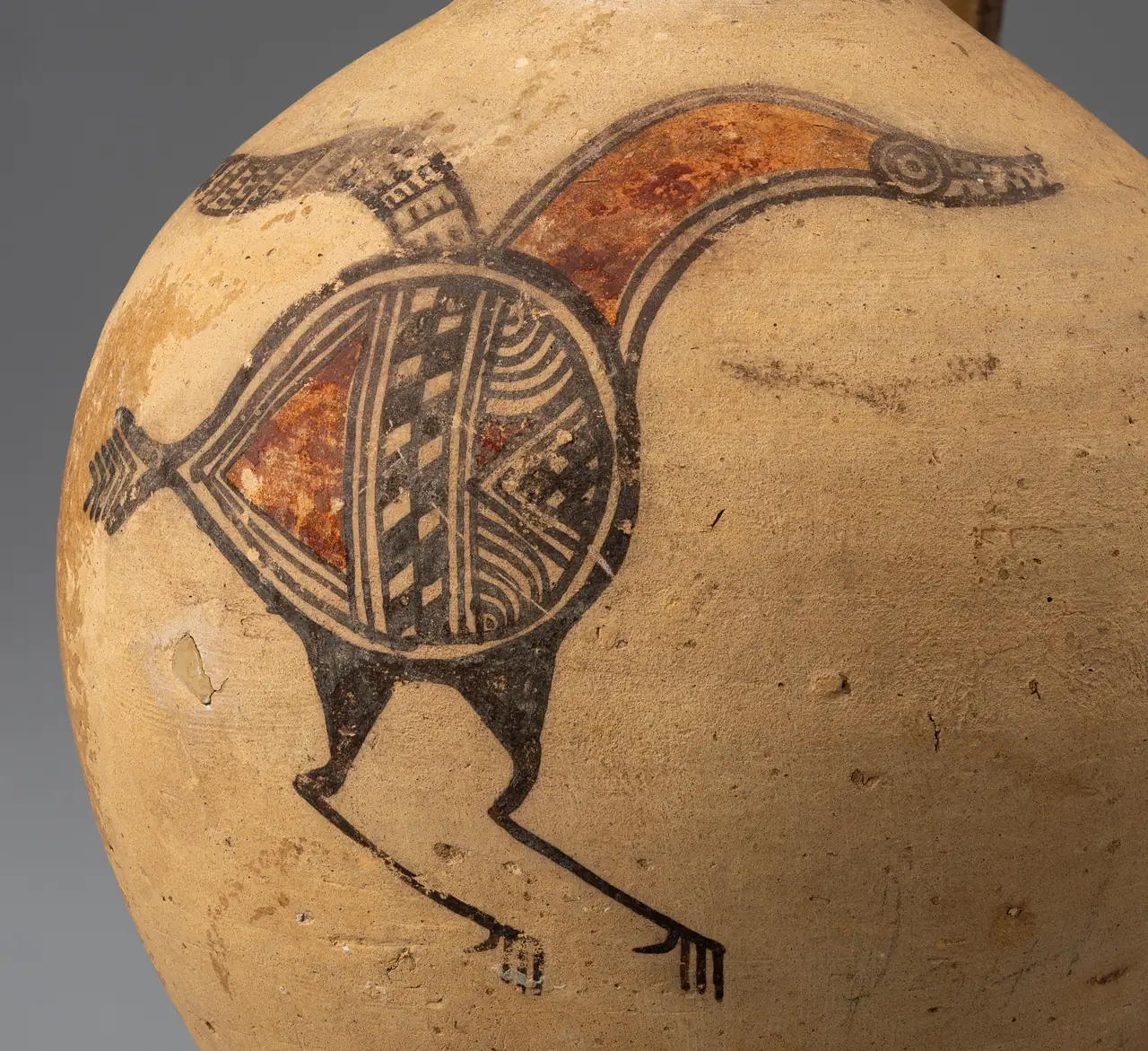 Close photograph of the painted bird on the jug: a round body divided into patterned segments, a red neck curving to a ringed eye and open beak, a wing plume, and long legs