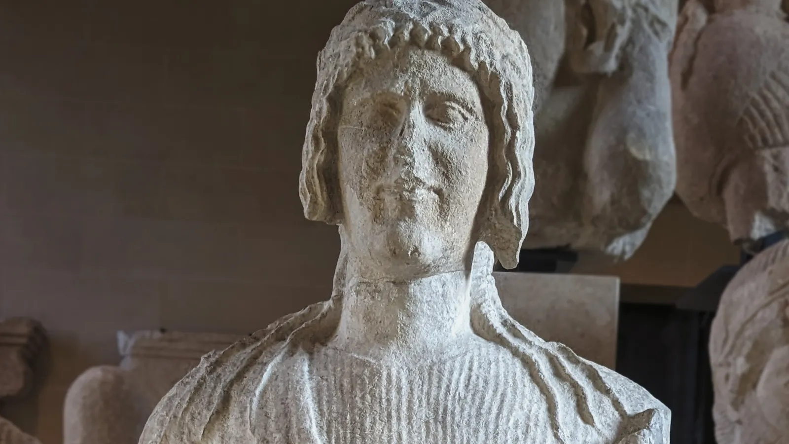 Head and shoulders of a weathered limestone statue of a young man with a beaded hair band and a pleated tunic, among other statues in a museum gallery