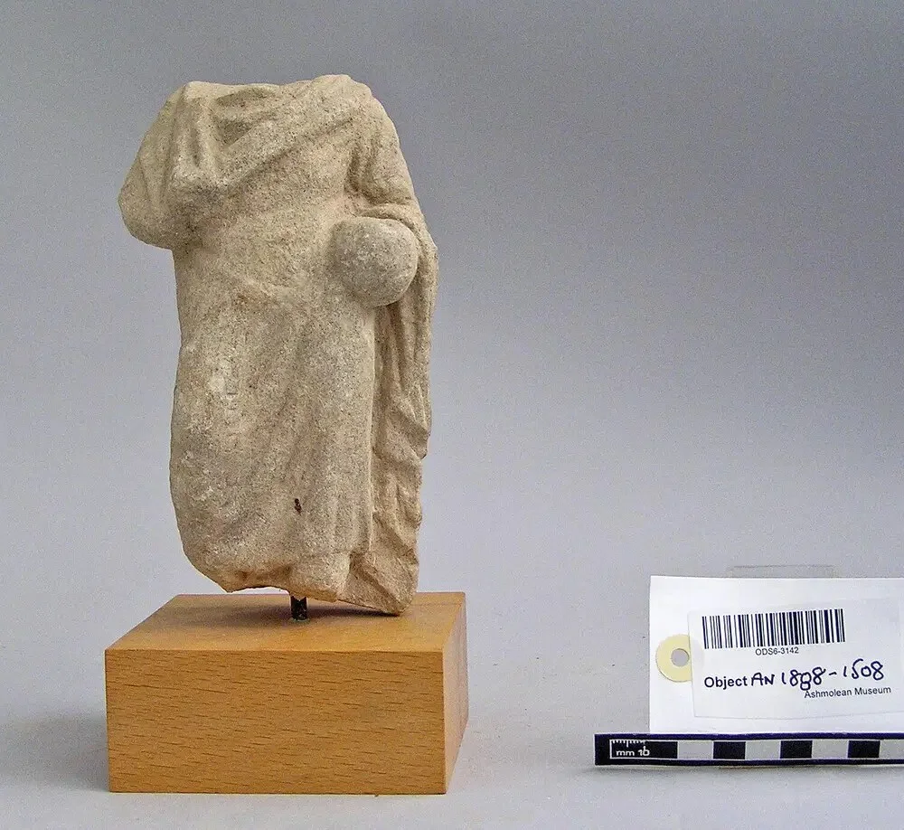 Headless limestone statuette of a draped standing figure holding a round object at its side, mounted on a wooden block beside a museum label and scale bar