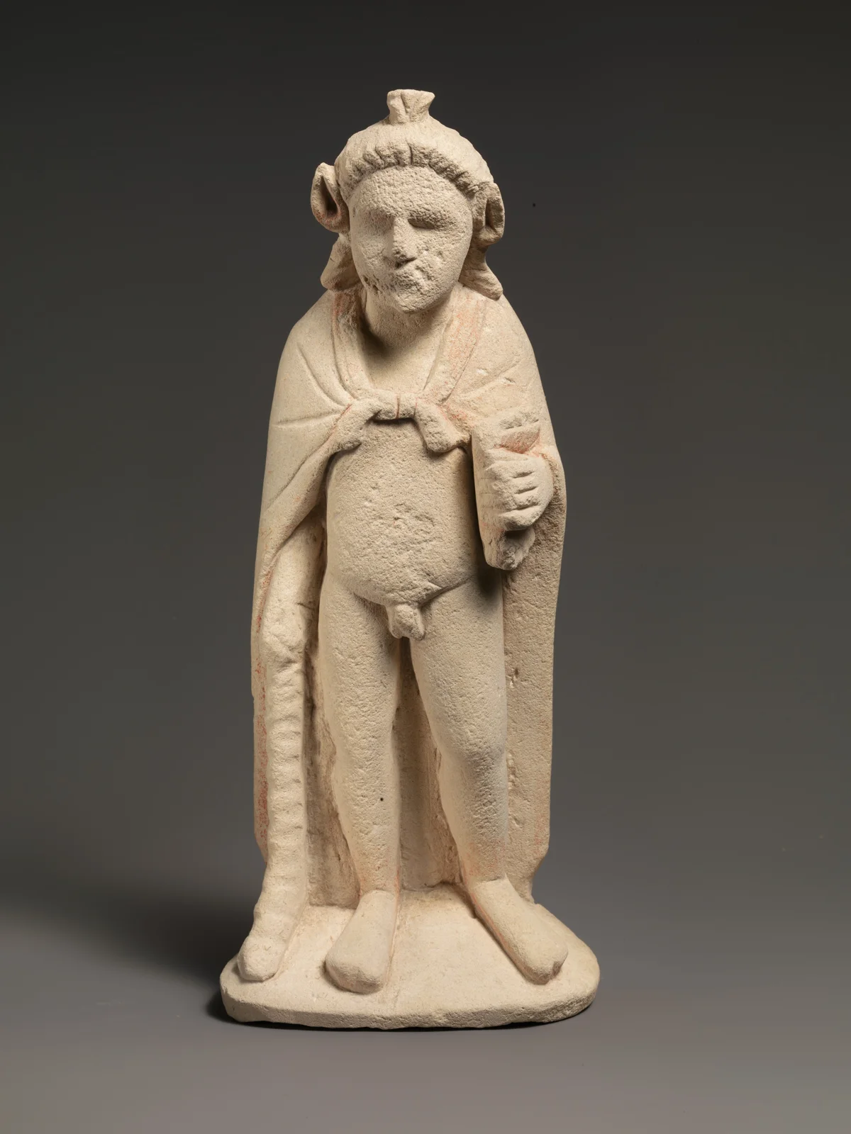 Limestone statuette of a bearded standing figure with a cloak knotted at the chest over a bare body, holding a staff at his side