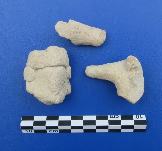 Three pale terracotta fragments on a blue background: a head with a headdress and round ear ornaments, and two broken limbs or bodies, beside a 10 cm scale