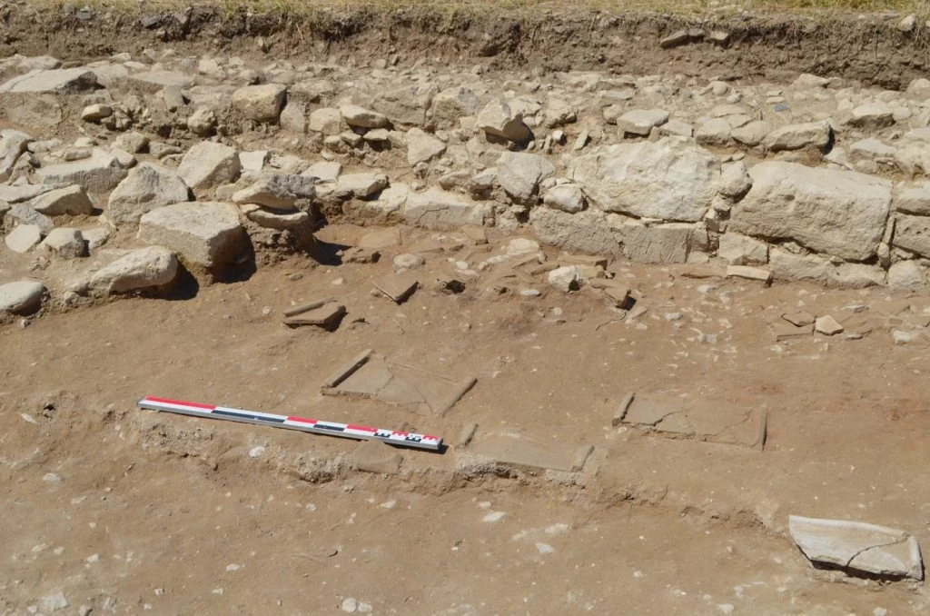 An excavated surface of reddish earth with a row of large pale limestone blocks along the back and flat broken tiles lying on the ground in front, with a red and white measuring rod