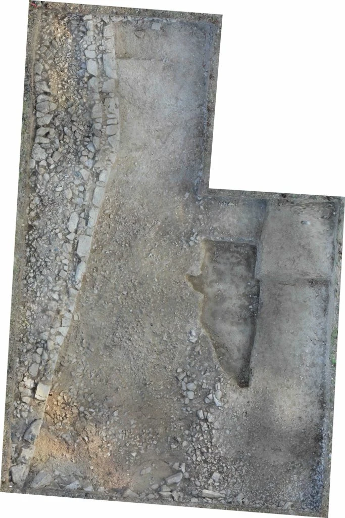 Overhead photograph of an L-shaped excavation trench, with a broad band of packed rough stones running down its left side and bare earth floors to the right