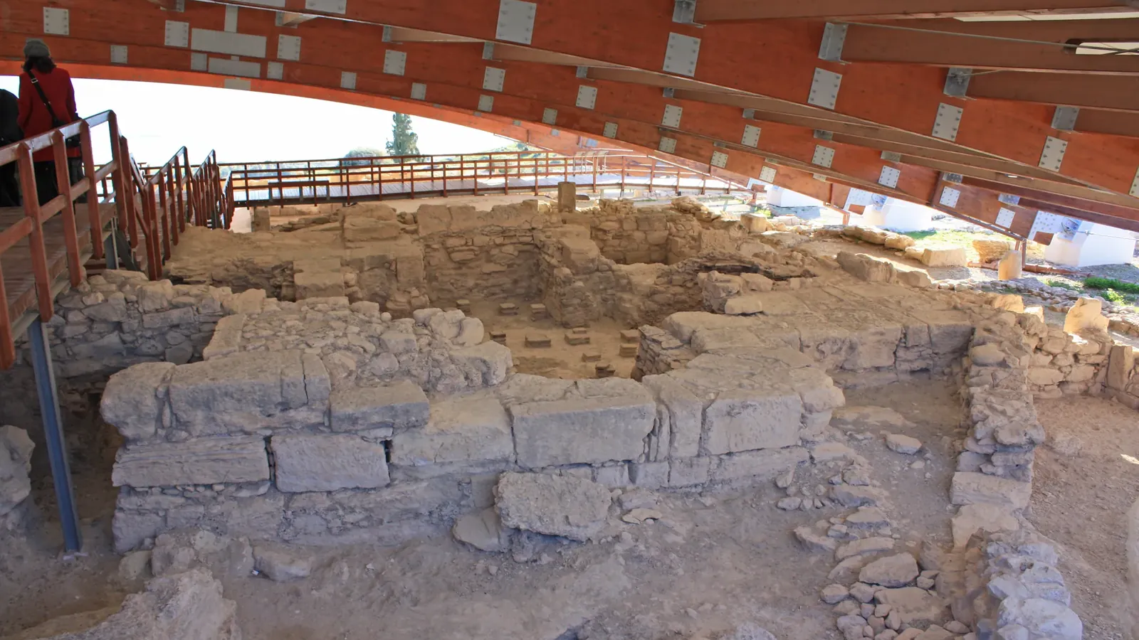 Excavated stone walls and rooms of an ancient house at Kourion under a curved timber roof, with a raised wooden walkway for visitors running along the far side