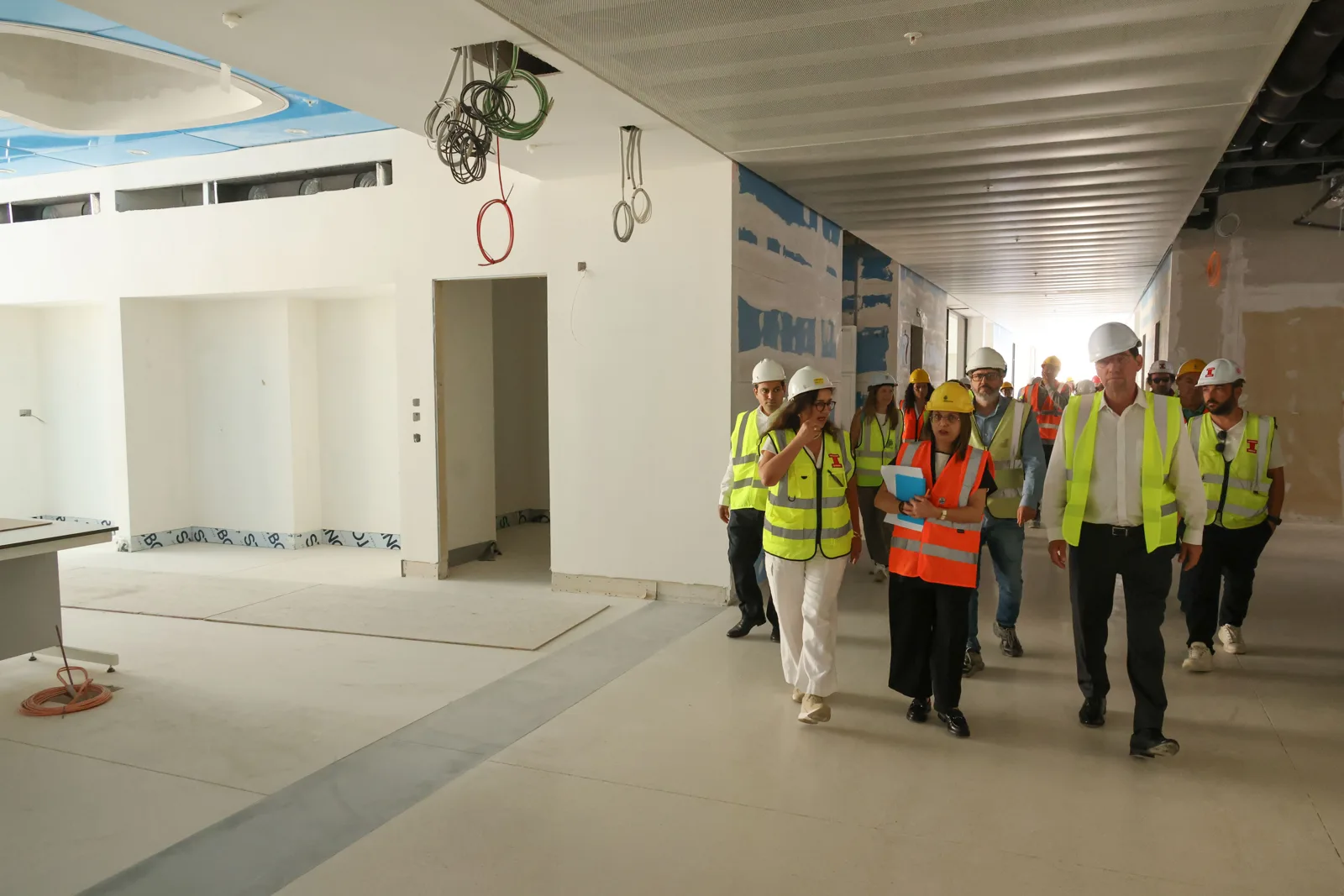 A group in hard hats and high-visibility vests walking down a wide, unfinished white corridor with ceiling services exposed