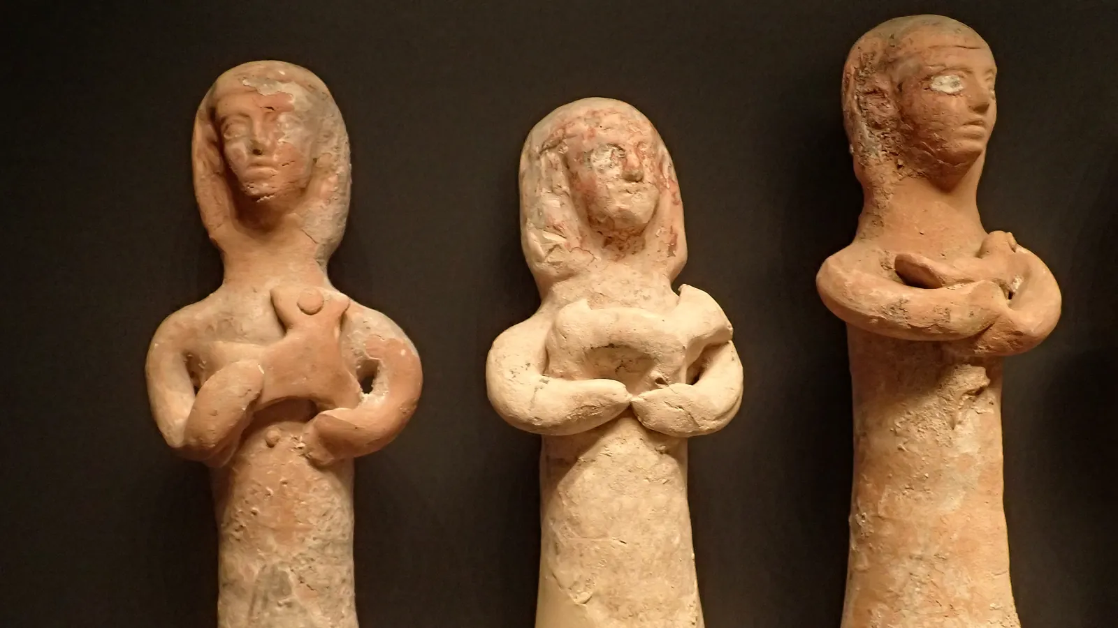 Three standing terracotta female figurines with covered heads, arms folded over objects held against their chests, in a museum case against a dark wall
