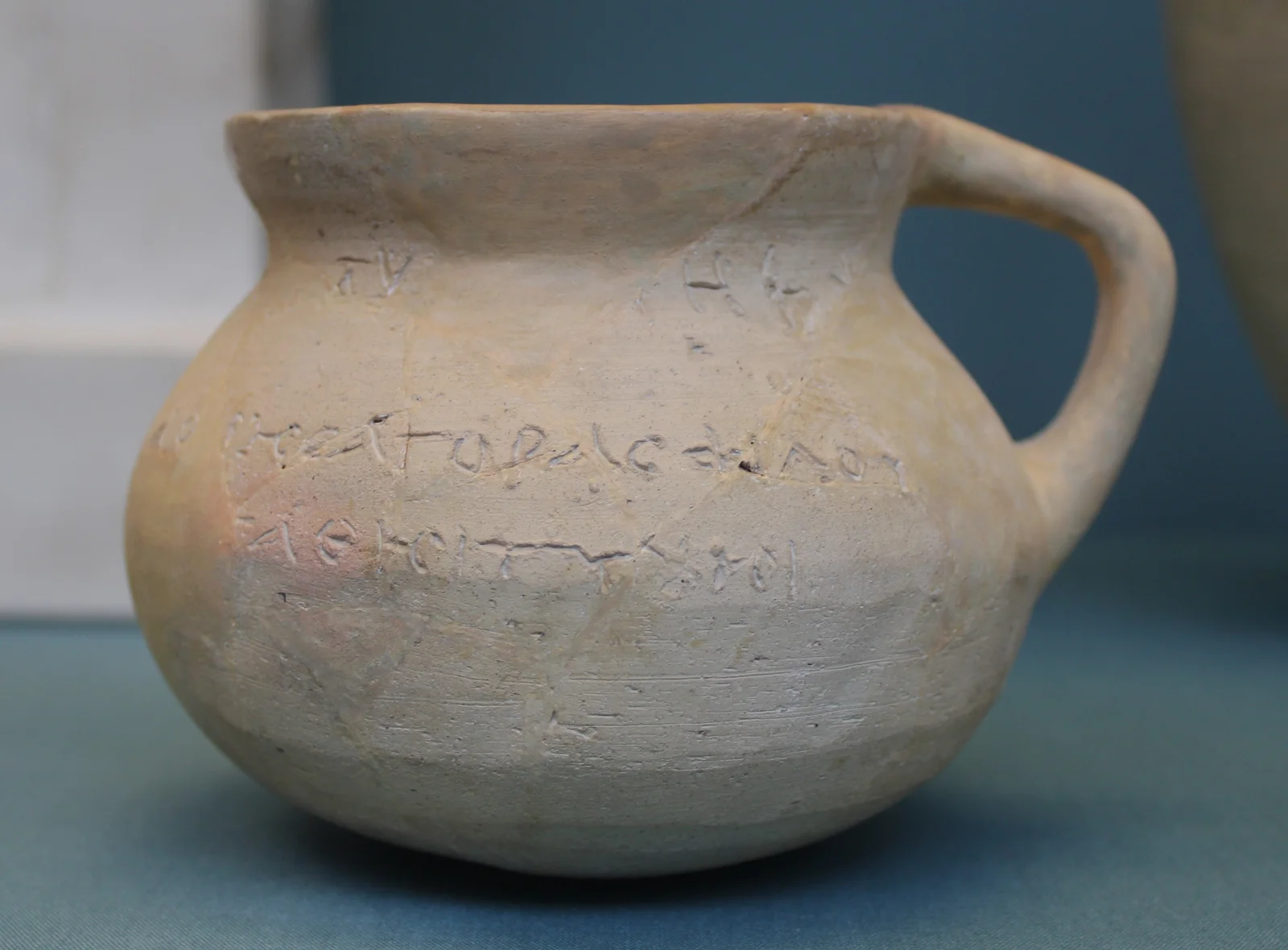 Small pale clay jug with one handle and a rounded body, with lines of scratched letters across the shoulder and belly