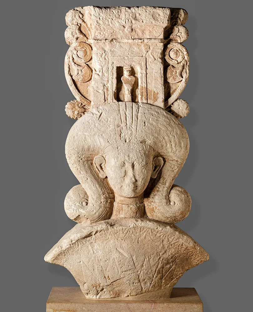Limestone capital carved with a woman's face framed by large curled locks, topped by a small shrine front with a figure in its doorway