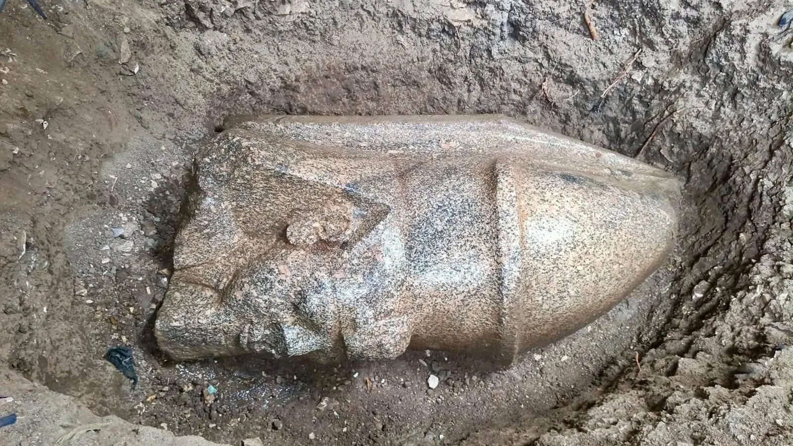 A granite head of a king wearing a tall crown, lying on its side at the bottom of a muddy excavation pit