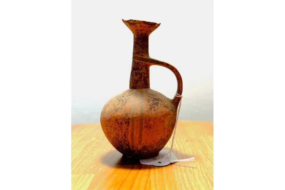 Small brown juglet with a round body, a tall narrow neck and a flared mouth, standing on a wooden table