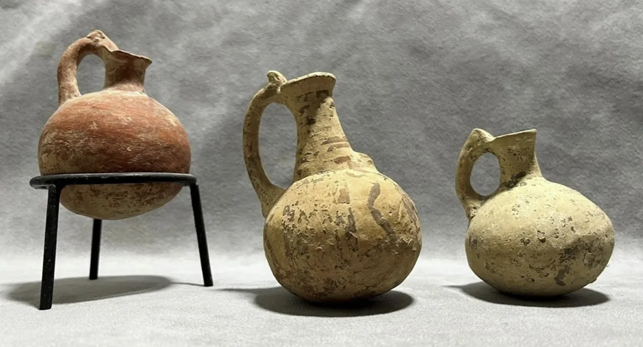 Three handmade pottery jugs on a grey cloth, the left one red and set in a black metal stand, the two on the right pale with dark painted bands