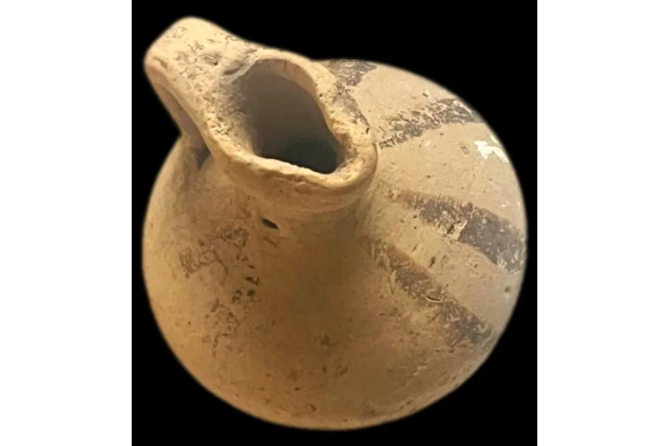 Close view of a pale clay juglet with a pinched spout and faded dark painted bands on the body, on a black background