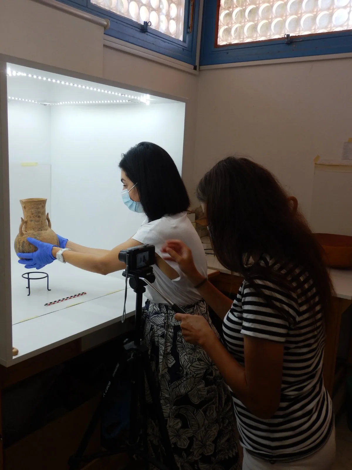 Two women in face masks and blue gloves photograph a two-handled clay jar standing alone in a lit glass display case