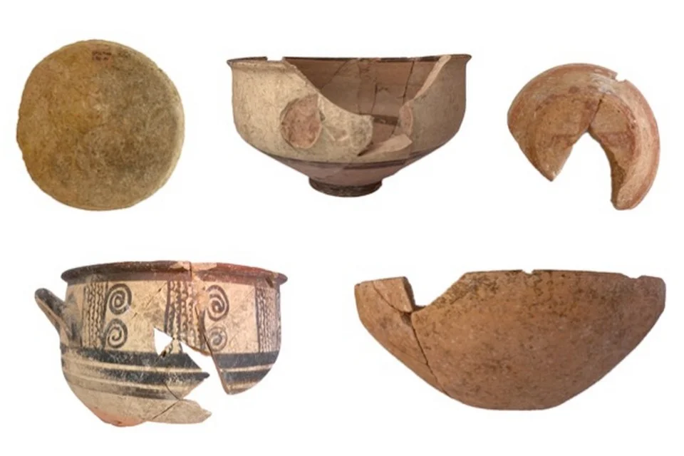 Five broken pottery vessels on a white ground: a round clay disc, a pale footed bowl, a curved fragment, a cup painted with black spirals and bands, and a plain deep bowl