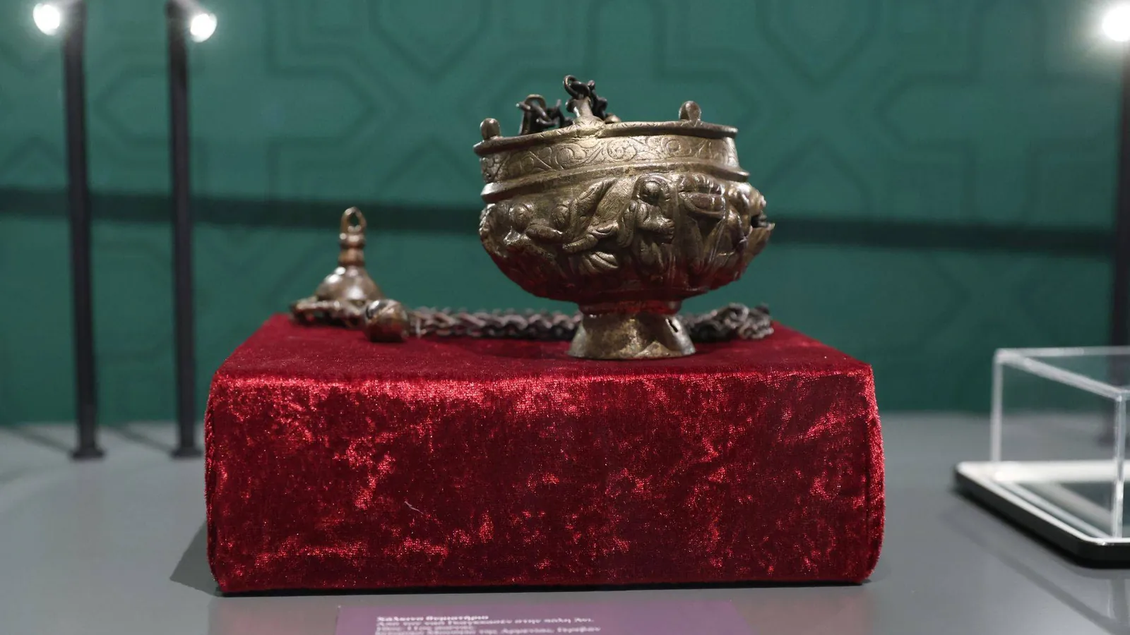 A bronze censer with a decorated bowl and its chains, resting on a red velvet stand in a museum display case