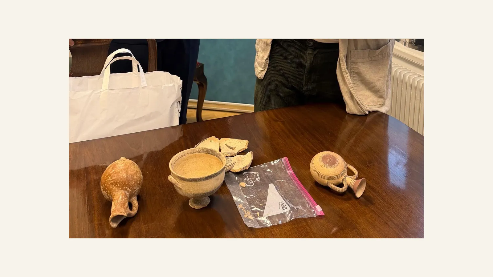 Pottery on a polished wooden table: a spouted jug and a small painted juglet lying on their sides, a two-handled bowl on a low foot, broken sherds and a plastic bag, with two people standing behind