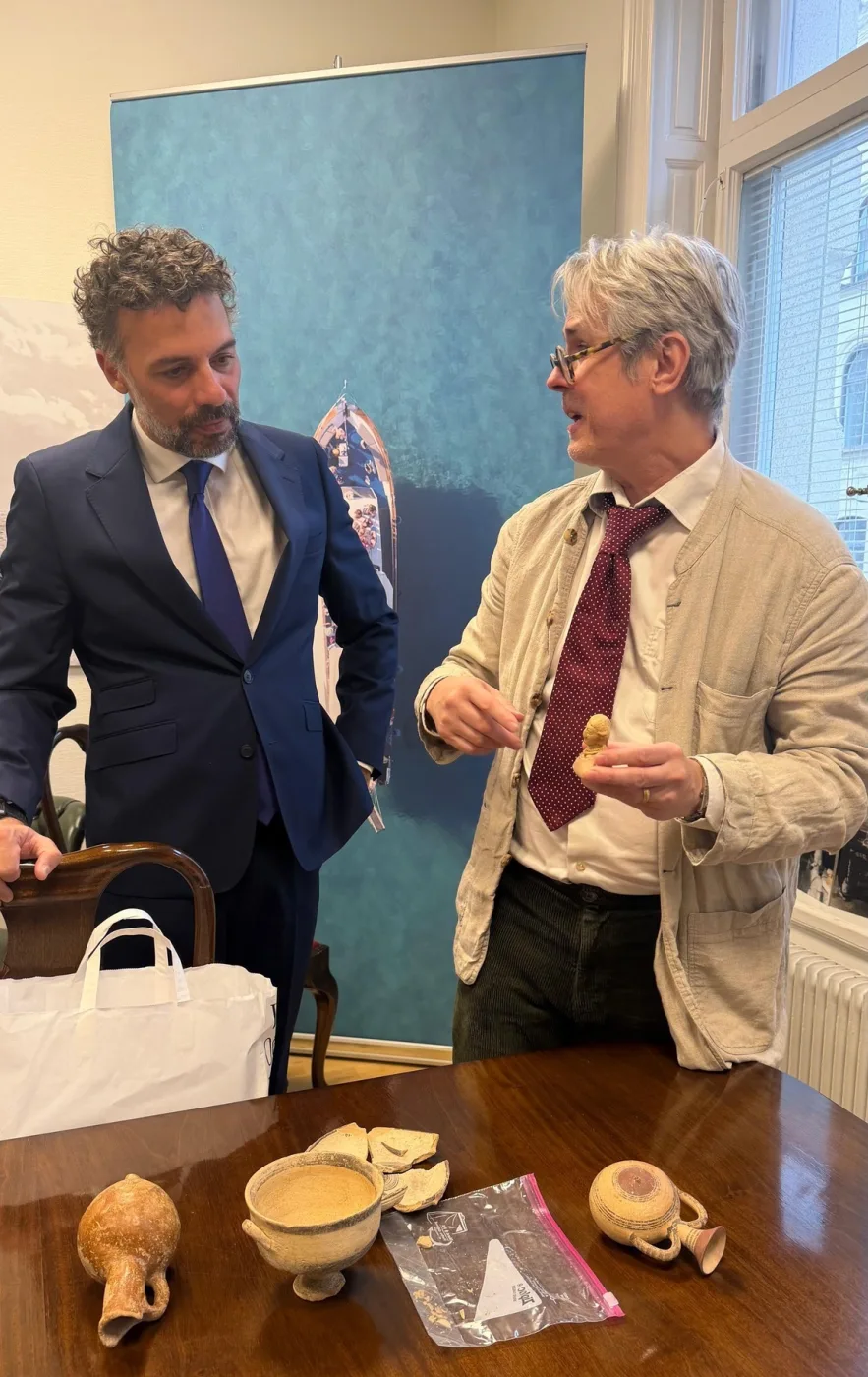 Two men in a meeting room beside a table with pottery on it; the man on the right, in a pale jacket and red tie, holds a small object in his hands while the man on the left, in a dark suit, looks on