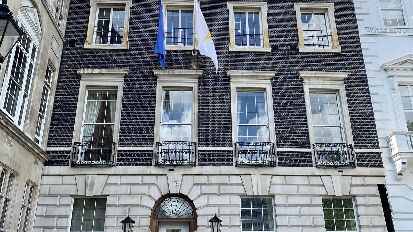 Dark brick Georgian town house with stone-framed sash windows and iron balconies, the EU and Cyprus flags above, and a fanlit door numbered 13 at street level