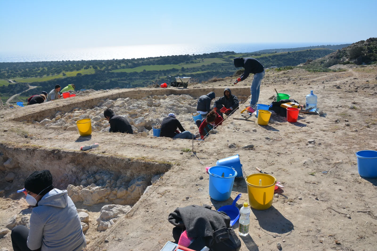 Excavators with buckets and trowels working in a shallow rectangular trench on a bare hilltop, with fields and the sea beyond
