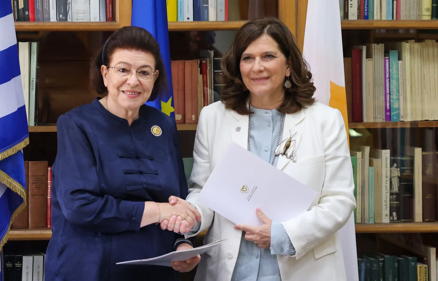 Two women shake hands in front of bookshelves and the Greek, EU and Cypriot flags; one wears a dark blue jacket, the other a white jacket and holds a white folder
