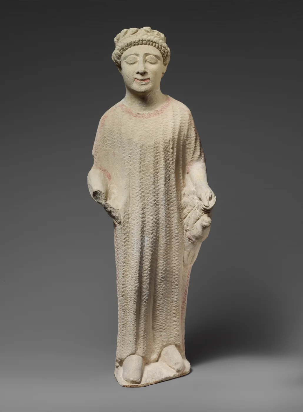 Limestone statuette of a standing boy in a long belted tunic with a wreath in his hair, holding a bird against his left side