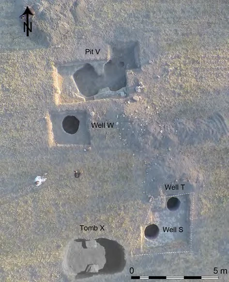 Aerial view of bare ground with five labelled cuttings: a large irregular pit at the top, three round well shafts and an oval tomb at the bottom, with a 5 m scale bar