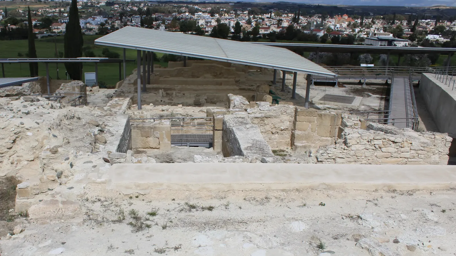 Excavated limestone walls and rooms on a hill, partly under modern metal shelters, with walkways and the houses of Dali on the plain beyond