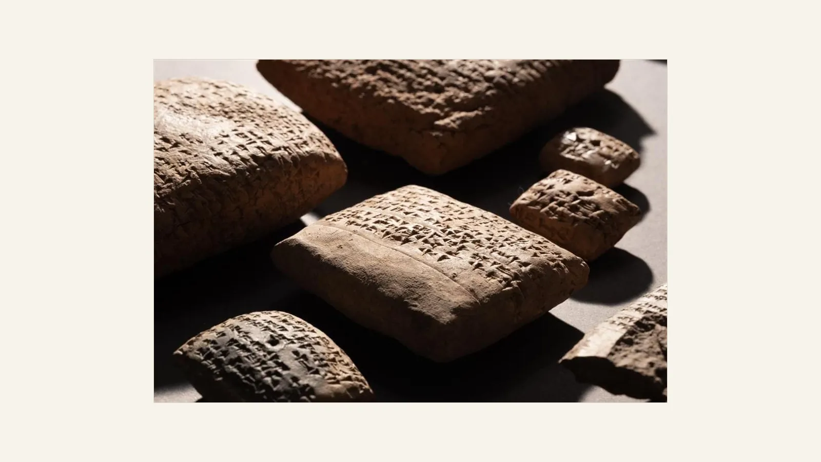 Several clay tablets densely impressed with cuneiform signs, lit from the side on a grey surface, with one rectangular tablet in focus at the centre