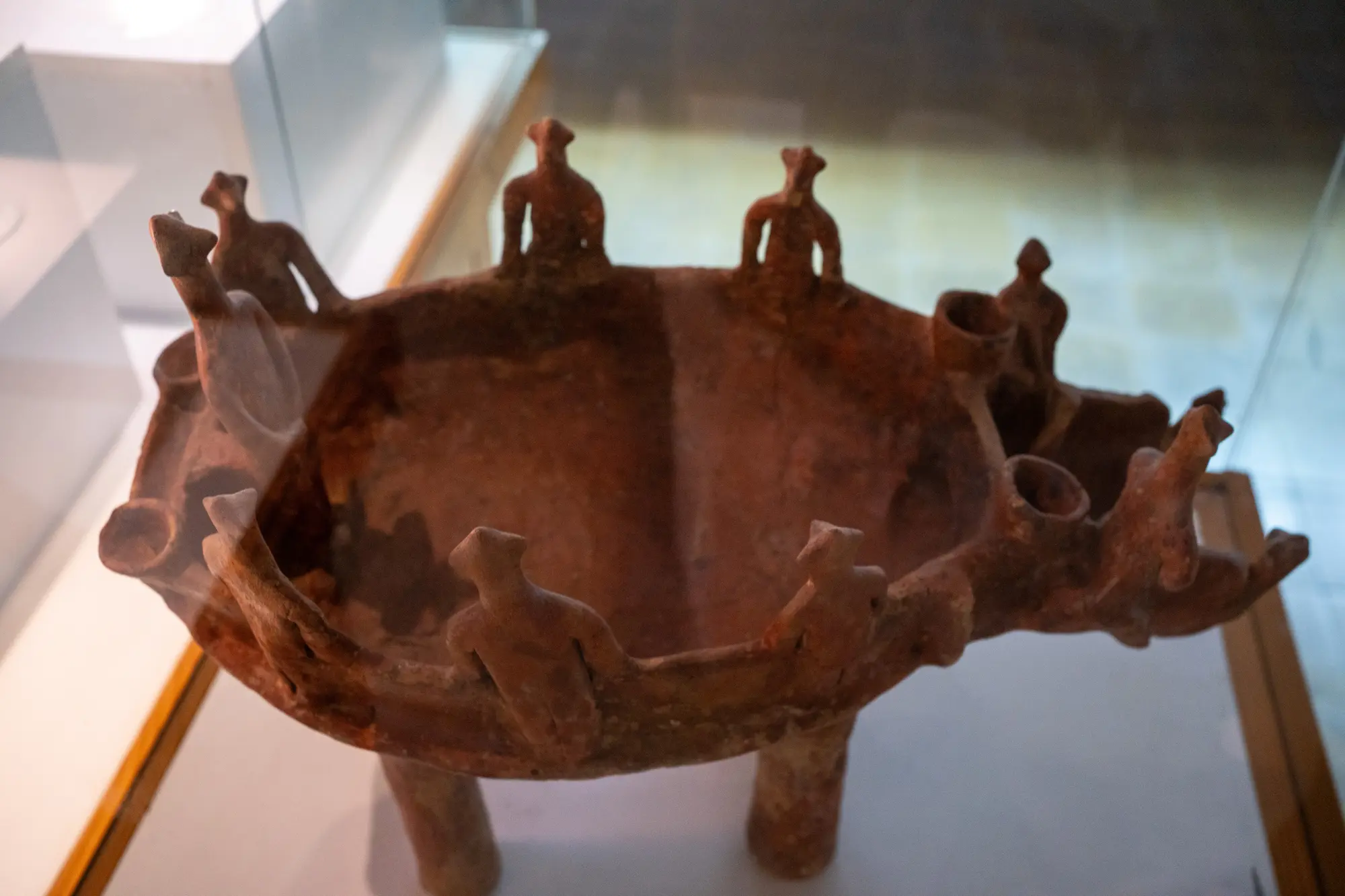 A red clay bowl on four legs with small modelled human figures and little jars set round its rim, in a glass case