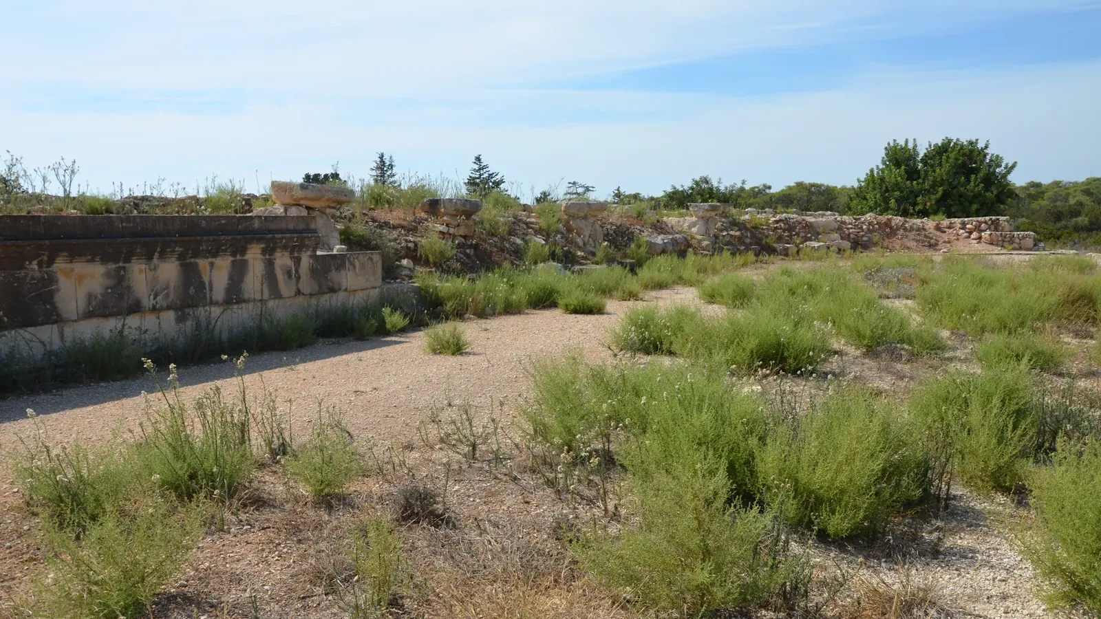 A gravel track with low scrub, edged on the left by a rebuilt bank of stone seating, with low ruined walls beyond