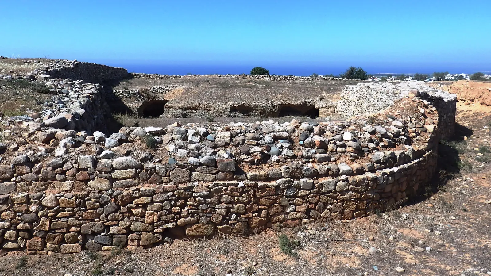 An earlier-excavated stretch of curved dry-stone city wall at the Northeast Gate on the Marcello hill, Kouklia, with open ground and the sea in the distance; the new tower is elsewhere on the wall