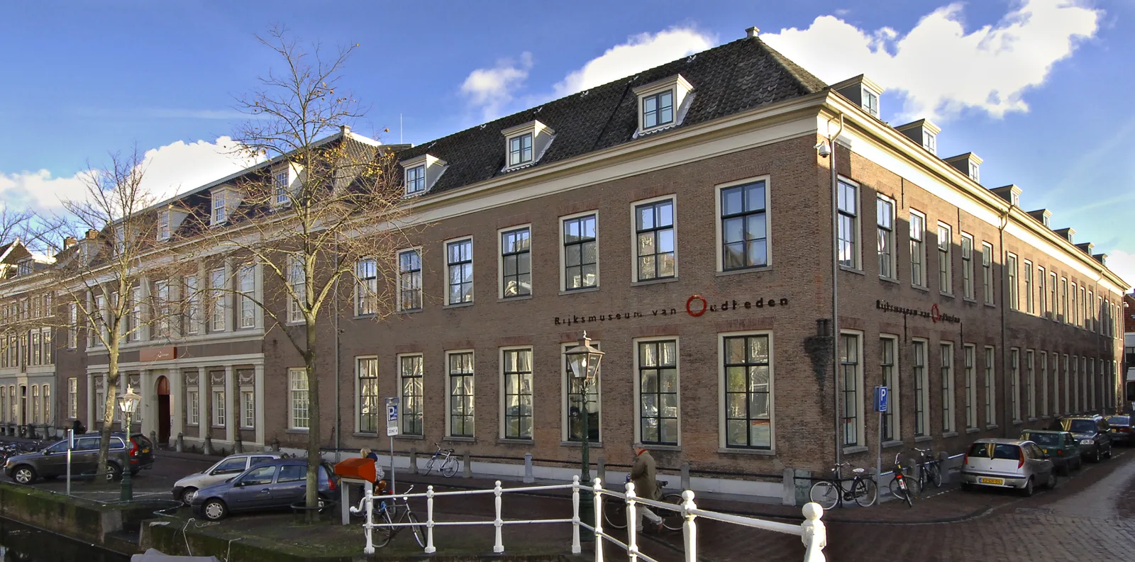A long three-storey brick building with rows of white-framed windows and dormers in the roof, on a street corner beside a canal with a white railing, bare trees and parked cars and bicycles