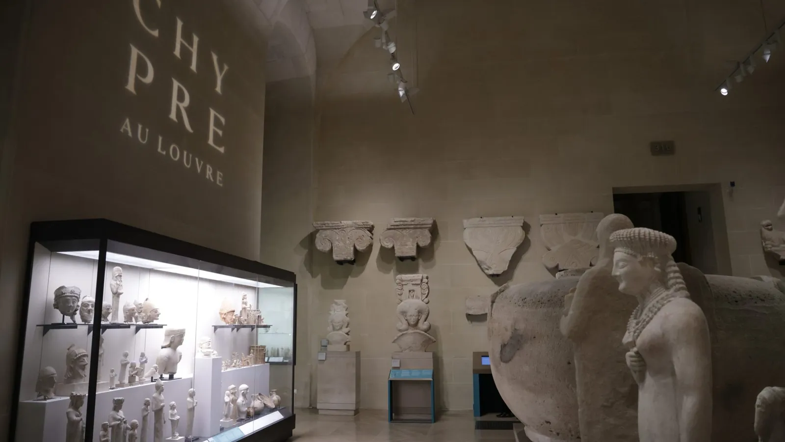 Museum gallery with a vaulted stone ceiling, a lit glass case of limestone heads and small figures on the left, carved stone capitals on the far wall, a large stone vase and two female statues on the right, and the words Chypre au Louvre projected on the wall