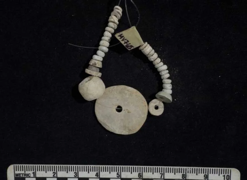 A short strand of small pale disc beads threaded on wire, with a large flat perforated disc, a round bead and a ring-shaped bead, beside a centimetre scale