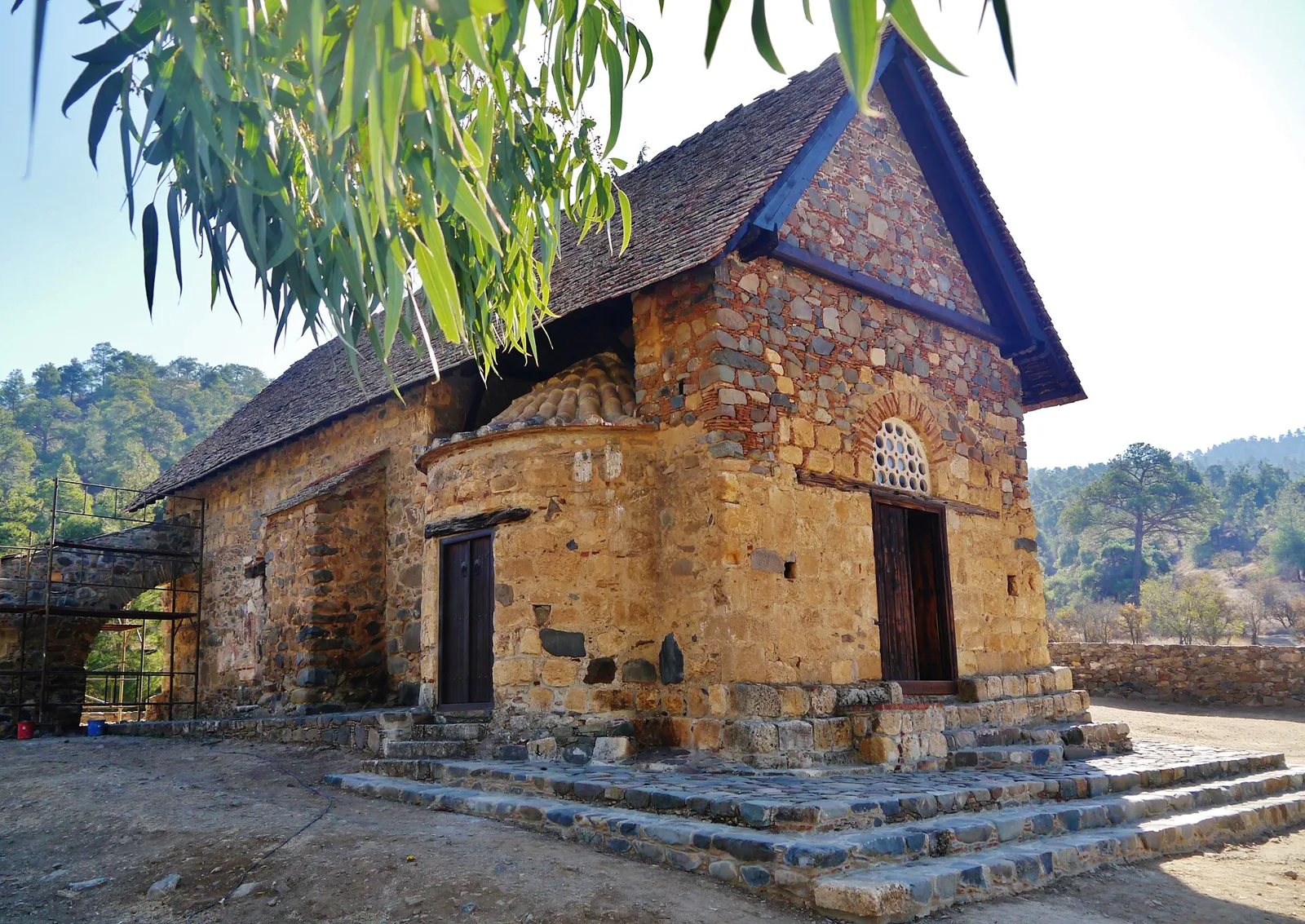 Small stone church with a steep pitched roof of flat tiles, a rounded apse and stone steps in front, among pine-covered hills