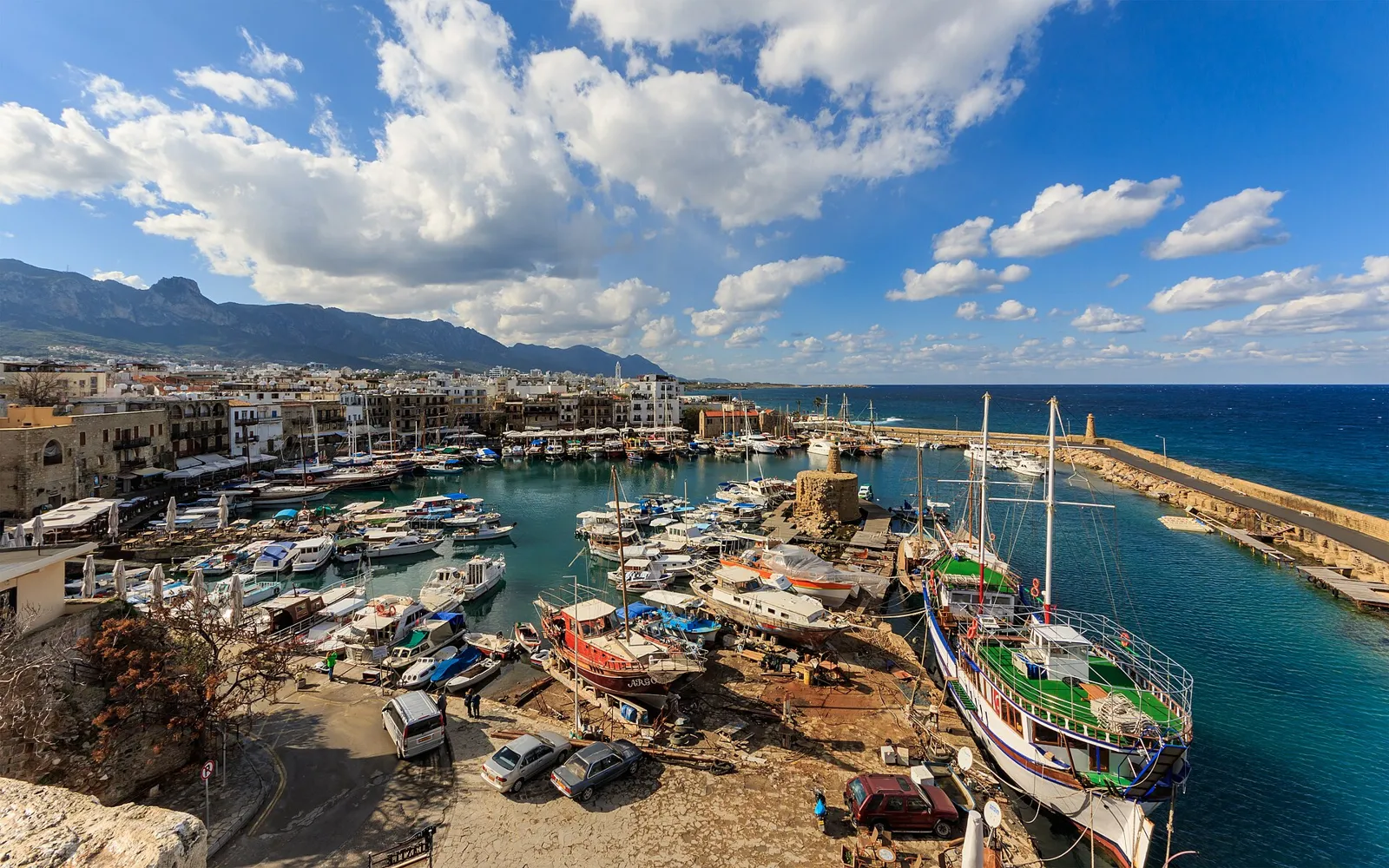 Horseshoe-shaped harbour crowded with fishing and tour boats, white town buildings behind it and the Kyrenia mountain range under clouds
